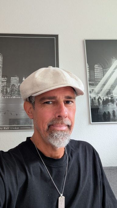 Mann mit grauem Bart und Mütze, lächelt vor einem modernen Hintergrund.
