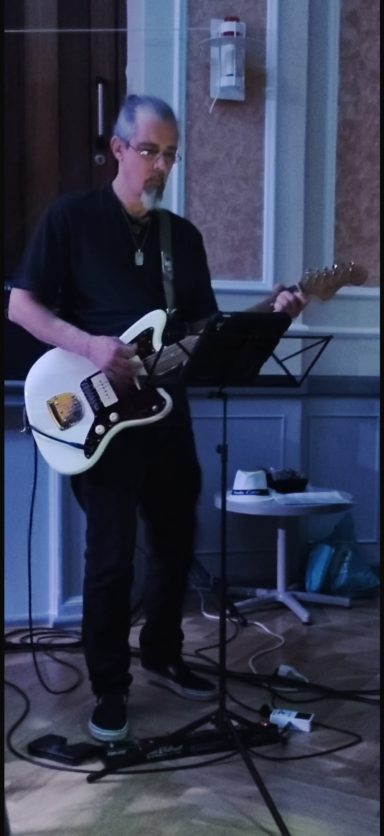 Mann spielt E-Gitarre auf einer Bühne, schwarz gekleidet mit Brille.