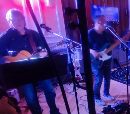 Zwei Musiker mit Gitarre und E-Gitarre, beleuchtet von bunten Lichtern auf der Bühne.