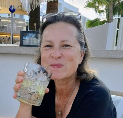 Frau mit einem Glas Getränk lächelt, im Freien, Sonnenlicht und Palmen im Hintergrund.